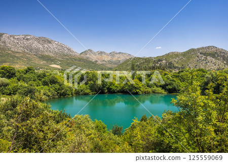 Modro Oko, small lake near delta of the Neretva river, Croatia, Europe. A serene turquoise lake nestled among lush green trees with rolling mountains under a clear blue sky, a peaceful nature scene. 125559069