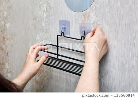 Person is pressing transparent adhesive hook onto bathroom wall while black metal shelf is already hanging on adjacent fixed hook. Person is pressing transparent adhesive hook onto bathroom wall while black metal shelf is already hanging on adjacent fixed hook. 125560914