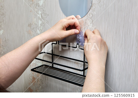 Hands are peeling off protective film from wall adhesive hook before securing black metal shelf to smooth vertical surface with patterned wallpaper. 125560915