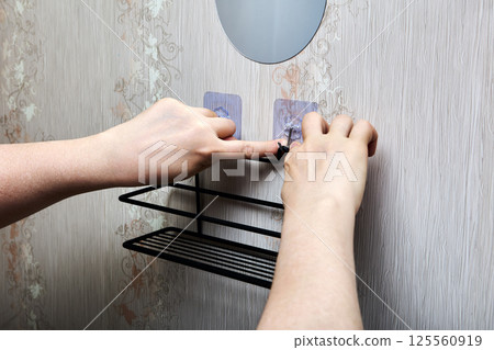 Hands press transparent peel-and-stick hook firmly against smooth plastic wall panel while black metal shelf is held in place. Hands press transparent peel-and-stick hook firmly against smooth plastic wall panel while black metal shelf is held in place. 125560919