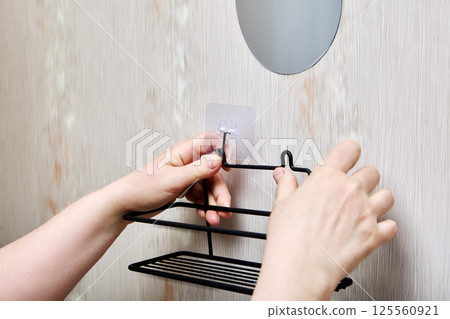 Person is attaching wire bathroom rack onto two fixed nano adhesive wall mounts ensuring correct placement by fitting hooks into mounting slots. 125560921