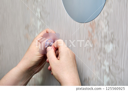Hands are removing protective film from nano adhesive hook before mounting it onto smooth plastic wall panel near round mirror. Hands are removing protective film from nano adhesive hook before mounting it onto smooth plastic wall panel near round mirror. 125560925