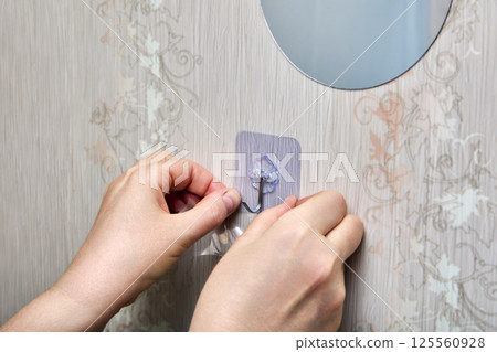 Person is pressing transparent adhesive patch with plastic hook onto vertical surface near round mirror, using both hands for secure attachment. Person is pressing transparent adhesive patch with plastic hook onto vertical surface near round mirror, using both hands for secure attachment. 125560928