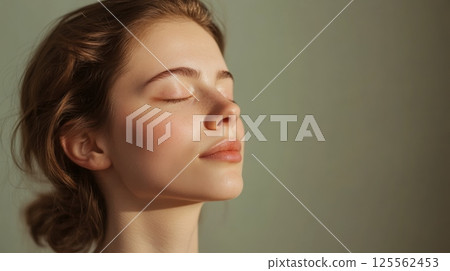 Serene young woman with closed eyes and a peaceful expression basks in the warm sunlight, showcasing healthy, glowing skin and a sense of tranquility 125562453