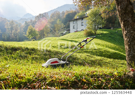 Lawn mower, brushcutter on green grass in garden in front of italian mountains, sunny autumn day. Valsassina 125563448