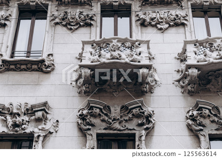 Ornate stone balcony and window frames with sculptural decorations on historic building facade. Architectural detail photograph. Baroque and Rococo influence. Urban architecture  125563614