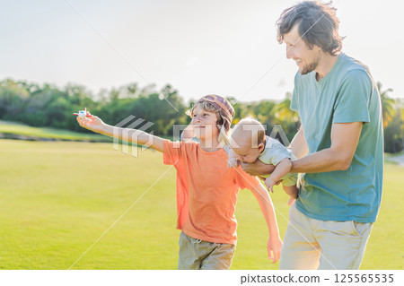 Father with his two sons in the park, the older boy wearing a pilot hat and holding a toy airplane while the baby watches. Childhood, bonding, and family adventure concept Father with his two sons in the park, the older boy wearing a pilot hat and holding a toy airplane while the baby watches. Childhood, bonding, and family adventure concept 125565535