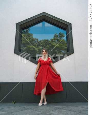 A woman stands elegantly in a flowing red dress, striking a pose in front of a unique hexagonal window. 125570296