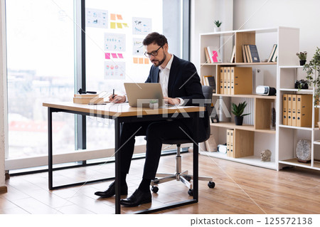 Young adult male wearing formal business attire seated in a modern office environment using a laptop and taking notes, showcasing productive work atmosphere. 125572138