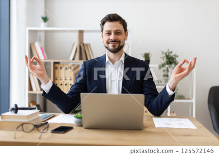 Professional adult male sitting at office desk in lotus pose meditating and smiling, embracing positive mindset in workplace environment for stress relief and cultivating mindfulness 125572146