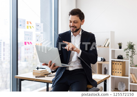 Professional adult Caucasian man wearing suit seated at desk holding laptop explaining concept Professional adult Caucasian man wearing suit seated at desk holding laptop explaining concept 125572150