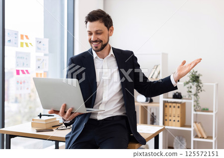 Young adult male business professional wearing suit sitting at desk conducting video meeting. Workplace setting includes books and plants. Confidence and enthusiasm displayed through gestures 125572151