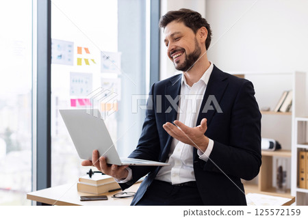 Professional adult Caucasian male in formal suit on video call speaking and gesturing at laptop, in bright modern office setting. Captures positive communication, digital technology, business concept. 125572159