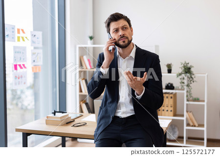 Middle-aged businessman in suit having phone call in bright office space, smiling with gesture, modern work environment 125572177