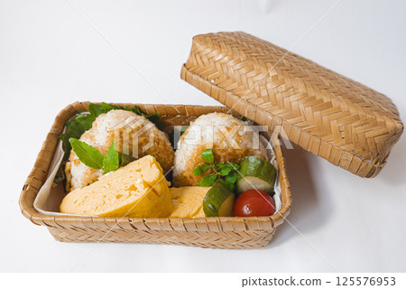 竹製便當盒裡放著以日本胡椒和芝麻烹調的味噌烤飯糰、煎蛋捲、米糠醃黃瓜等,背景則是白色的一張照片。 竹製便當盒裡放著以日本胡椒和芝麻烹調的味噌烤飯糰、煎蛋捲、米糠醃黃瓜等,背景則是白色的一張照片。 125576953