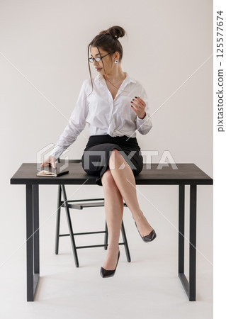 Professional businesswoman sitting on desk with document in hand, looking down at papers in modern office setting Professional businesswoman sitting on desk with document in hand, looking down at papers in modern office setting 125577674