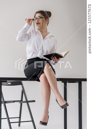 Focused young professional woman sitting on chair with book in hand, adjusting glasses Focused young professional woman sitting on chair with book in hand, adjusting glasses 125577678