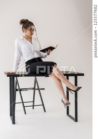 Woman in business suit sitting on desk, reading a book in white room Woman in business suit sitting on desk, reading a book in white room 125577682