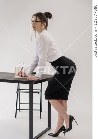 Attractive businesswoman in black pencil skirt and white shirt leaning on desk while writing in notebook Attractive businesswoman in black pencil skirt and white shirt leaning on desk while writing in notebook 125577686