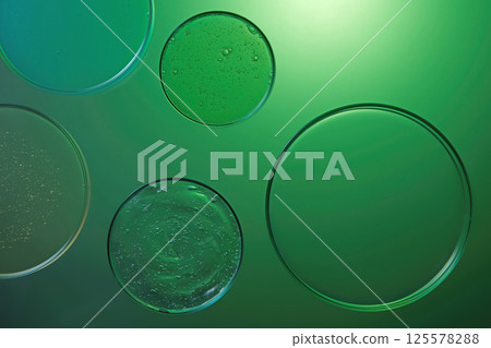 Petri dishes containing cosmetic essence, of various sizes. The essence has a thick texture with air bubbles. Scattered around on a green liquid background, light shining on the upper of the photo 125578288