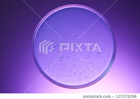 Round glass dish containing cosmetic liquid prepared in the laboratory. The air bubbles inside the water are small and dense. The background has a gradient effect from dark purple to light purple. 125578296