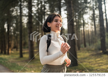 Athletic woman in a white outfit and black running backpack jogging through lush forest trails, blending fitness and adventure vibes 125579135