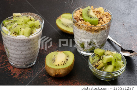 Two glasses of chia seed pudding, one topped with diced kiwi and the other with granola and banana chips, are on a dark surface with ingredients Two glasses of chia seed pudding, one topped with diced kiwi and the other with granola and banana chips, are on a dark surface with ingredients 125581287