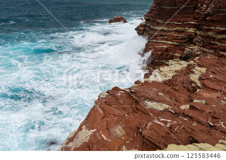 Red rock cliff in Cala Rossa, Terrasini, coastal waves crashing against rugged cliffs during daylight 125583446