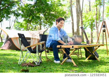 Young man using laptop at a table while camping in the forest Young man using laptop at a table while camping in the forest 125583451