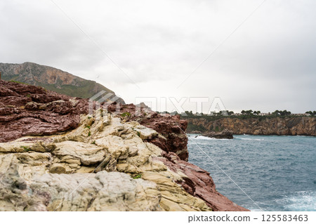 Red rock cliff in Cala Rossa, Terrasini, coastal landscape with rocky cliffs and ocean waves o cloudy day Red rock cliff in Cala Rossa, Terrasini, coastal landscape with rocky cliffs and ocean waves o cloudy day 125583463