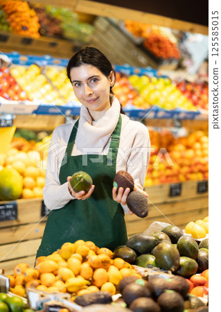 Young woman seller offering avocado in vegetable shop 125585015