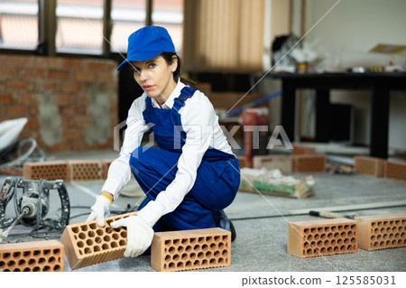 Woman builder in blue overalls lays out bricks on the wall in room 125585031