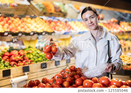 Woman carefully selects fresh tomatoes, standing near the counter in supermarket Woman carefully selects fresh tomatoes, standing near the counter in supermarket 125585046