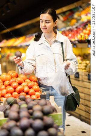 Woman carefully selects fresh tomatoes, standing near the counter in supermarket 125585107