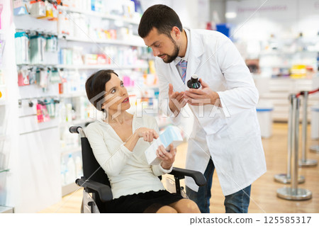 Male pharmacist consulting a woman in a wheelchair in pharmacy Male pharmacist consulting a woman in a wheelchair in pharmacy 125585317