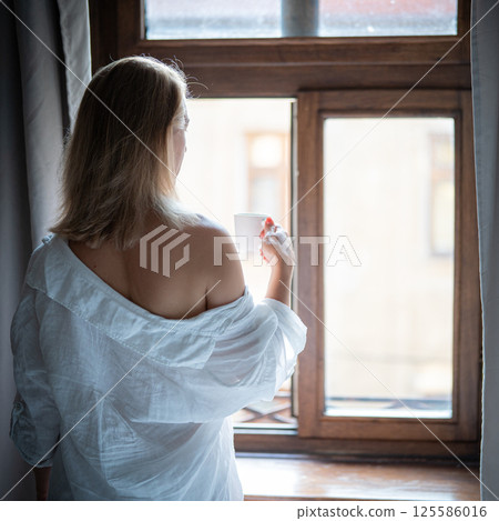 Woman in elegant robe drinking coffee in hotel room and standing near window 125586016