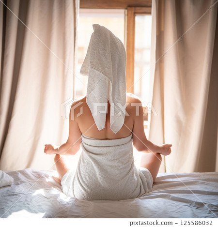 Back view of young woman in towel sitting on bed and meditating 125586032