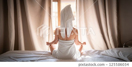 Back view of young woman in towel sitting on bed and meditating 125586034