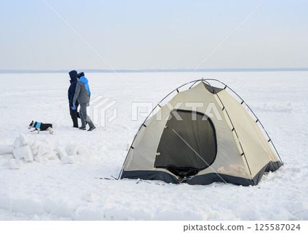 A tent in the snow, walking man, woman and a dog in the distance on a large plain of a frozen lake in winter A tent in the snow, walking man, woman and a dog in the distance on a large plain of a frozen lake in winter 125587024