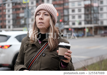Portrait of mature woman in wool hat standing on the urban street, drinking takeaway coffee from paper cup. Middle-aged 40s female walking around the city at cold autumn day. Living in modern district Portrait of mature woman in wool hat standing on the urban street, drinking takeaway coffee from paper cup. Middle-aged 40s female walking around the city at cold autumn day. Living in modern district 125587089