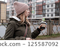 Portrait of mature woman in wool hat standing on the urban street, drinking takeaway coffee from paper cup. Middle-aged 40s female walking around the city at cold autumn day. Living in modern district 125587092