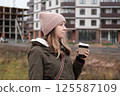 Portrait of mature woman in wool hat standing on the urban street, drinking takeaway coffee from paper cup. Middle-aged 40s female walking around the city at cold autumn day. Living in modern district 125587109