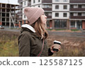 Portrait of mature woman in wool hat standing on the urban street, drinking takeaway coffee from paper cup. Middle-aged 40s female walking around the city at cold autumn day. Living in modern district 125587125