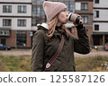 Portrait of mature woman in wool hat standing on the urban street, drinking takeaway coffee from paper cup. Middle-aged 40s female walking around the city at cold autumn day. Living in modern district 125587126