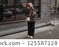 Portrait of mature woman in wool hat standing on the urban street, drinking takeaway coffee from paper cup. Middle-aged 40s female walking around the city at cold autumn day. Living in modern district 125587128