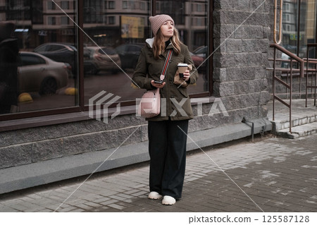 Portrait of mature woman in wool hat standing on the urban street, drinking takeaway coffee from paper cup. Middle-aged 40s female walking around the city at cold autumn day. Living in modern district Portrait of mature woman in wool hat standing on the urban street, drinking takeaway coffee from paper cup. Middle-aged 40s female walking around the city at cold autumn day. Living in modern district 125587128