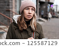 Portrait of mature woman in wool hat standing on the urban street, drinking takeaway coffee from paper cup. Middle-aged 40s female walking around the city at cold autumn day. Living in modern district 125587129