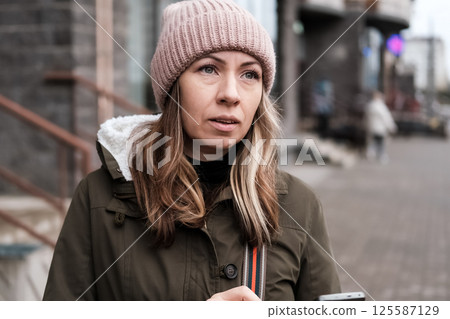 Portrait of mature woman in wool hat standing on the urban street, drinking takeaway coffee from paper cup. Middle-aged 40s female walking around the city at cold autumn day. Living in modern district Portrait of mature woman in wool hat standing on the urban street, drinking takeaway coffee from paper cup. Middle-aged 40s female walking around the city at cold autumn day. Living in modern district 125587129