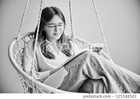 Beautiful girl relaxing and reading book on indoor swing. Pretty female in glasses excited about story she reading. Cozy afternoon with book in hanging chair in home. Black and white image. 125588501