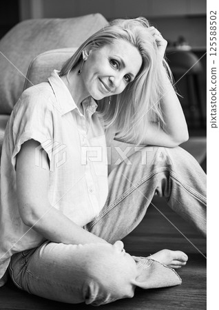 Portrait of pretty woman with blonde hair relaxing next to couch. Beautiful woman sitting on wooden floor. Black and white image. 125588502